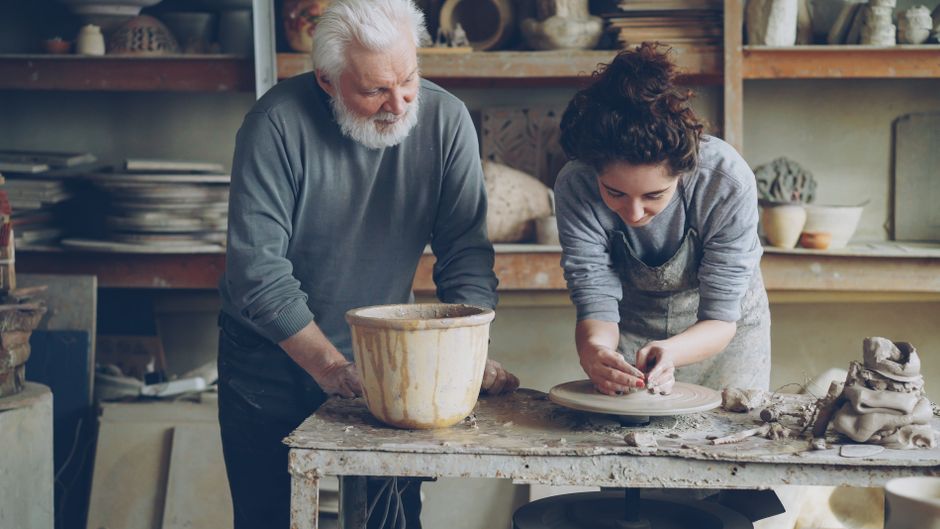 Elderly man teaching woman pottery in a cozy studio setting, focusing on skill development.