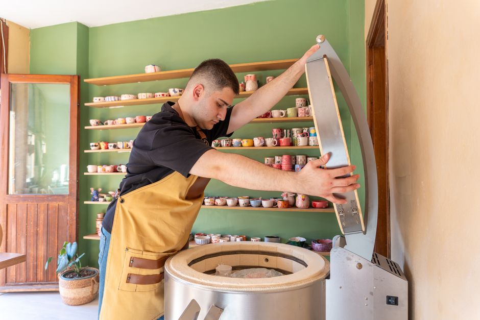 A man in a pottery studio opening a kiln surrounded by colorful ceramics on shelves.